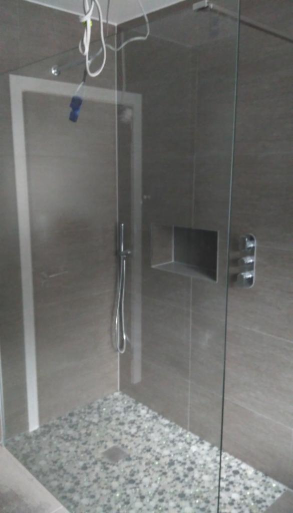 Nowoczesna kabina prysznicowa z szarą glazurą, brodzikiem wyłożonym drobnymi kamieniami i szklaną ścianką, widoczne chromowane elementy armatury i wnęka w ścianie.