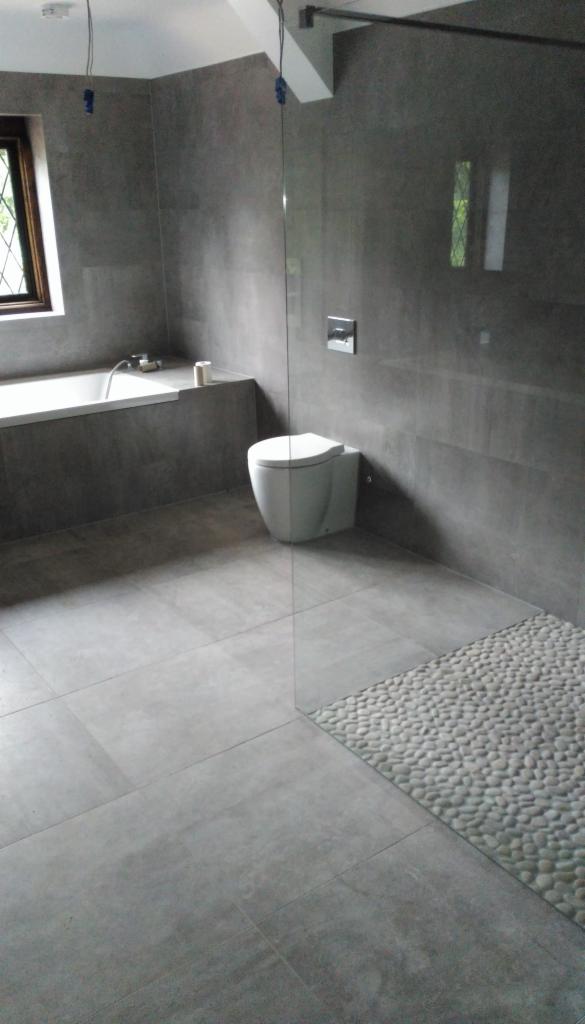 Nowoczesna łazienka z szarymi płytkami wielkoformatowymi na ścianach i podłodze, kabina prysznicowa ze szklaną ścianką, fragment podłogi wyłożony drobnymi, jasnymi kamieniami, wisząca toaleta...