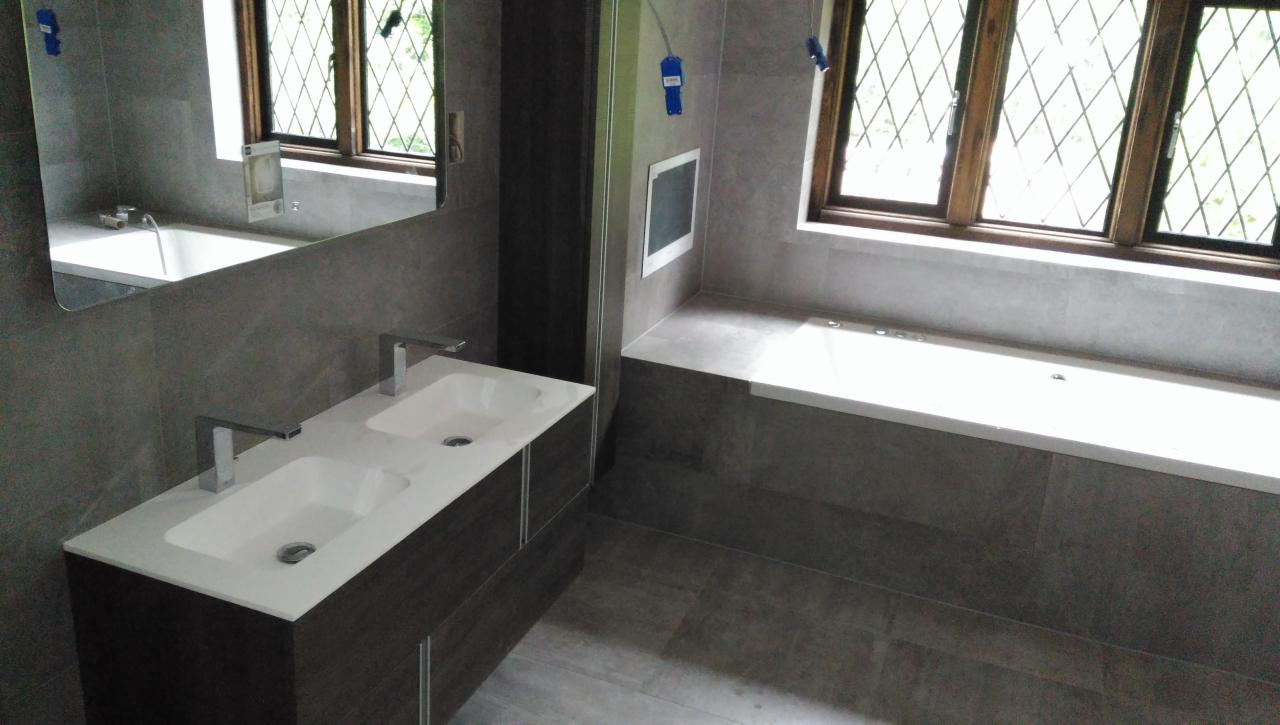 Nowoczesna łazienka z dwoma umywalkami na szafce z ciemnego drewna, szare płytki, okno z kratownicą i zabudowana wanna.