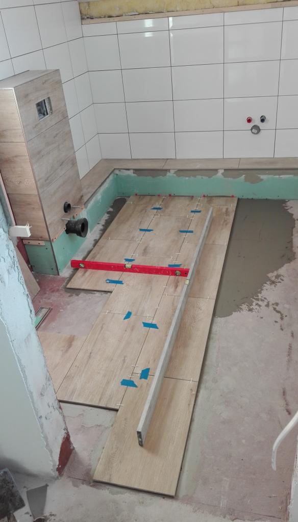 Układanie płytek podłogowych imitujących drewno w łazience, poziomica na płytkach, widoczne krzyżyki dystansowe i niebieska taśma klejąca, białe płytki na ścianach z widocznymi przyłączami wodnymi.