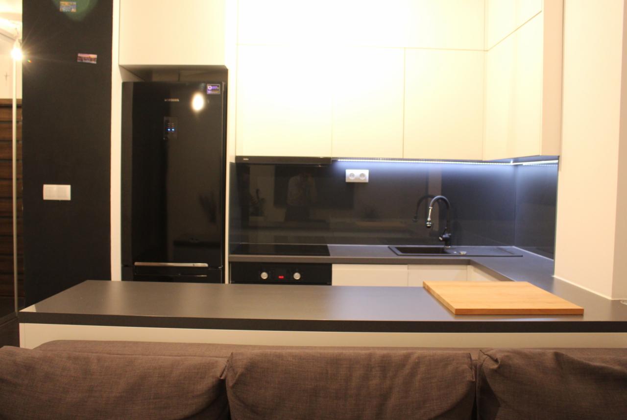 Nowoczesna kuchnia z białymi szafkami, czarnym blatem i panelem szklanym, oświetleniem LED pod szafkami, widoczna deska do krojenia i fragment sofy w kolorze brązowym.