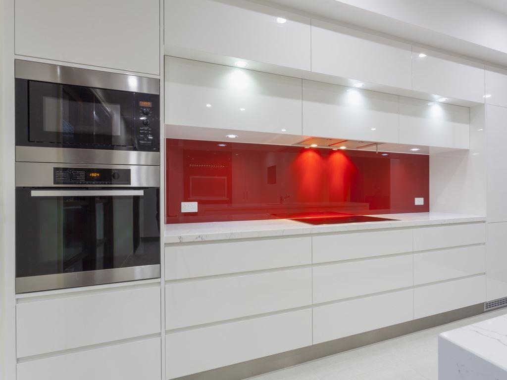 Biała, nowoczesna kuchnia z połyskiem, czerwony panel szklany nad blatem, wbudowany piekarnik i mikrofalówka.