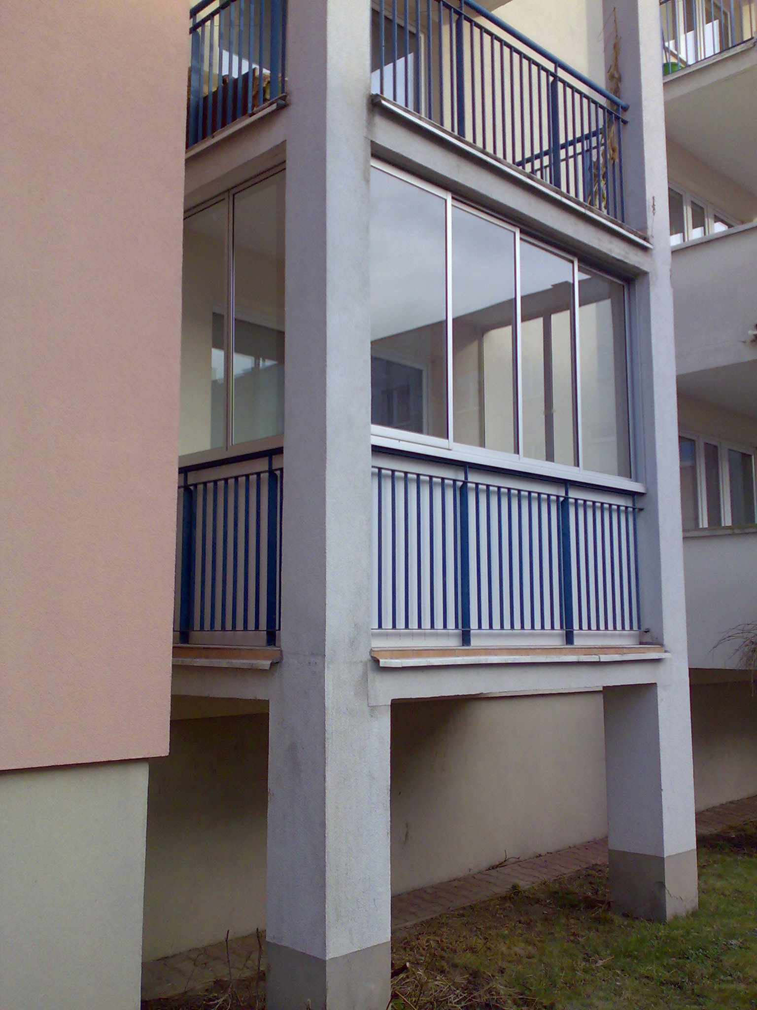 Zabudowany balkon z aluminiowymi ramami okiennymi, niebieska metalowa balustrada, widok z zewnątrz na dwukondygnacyjny budynek.