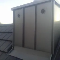 Szary komin wentylacyjny z dwoma okrągłymi kratkami na dachu pokrytym ciemną dachówką.