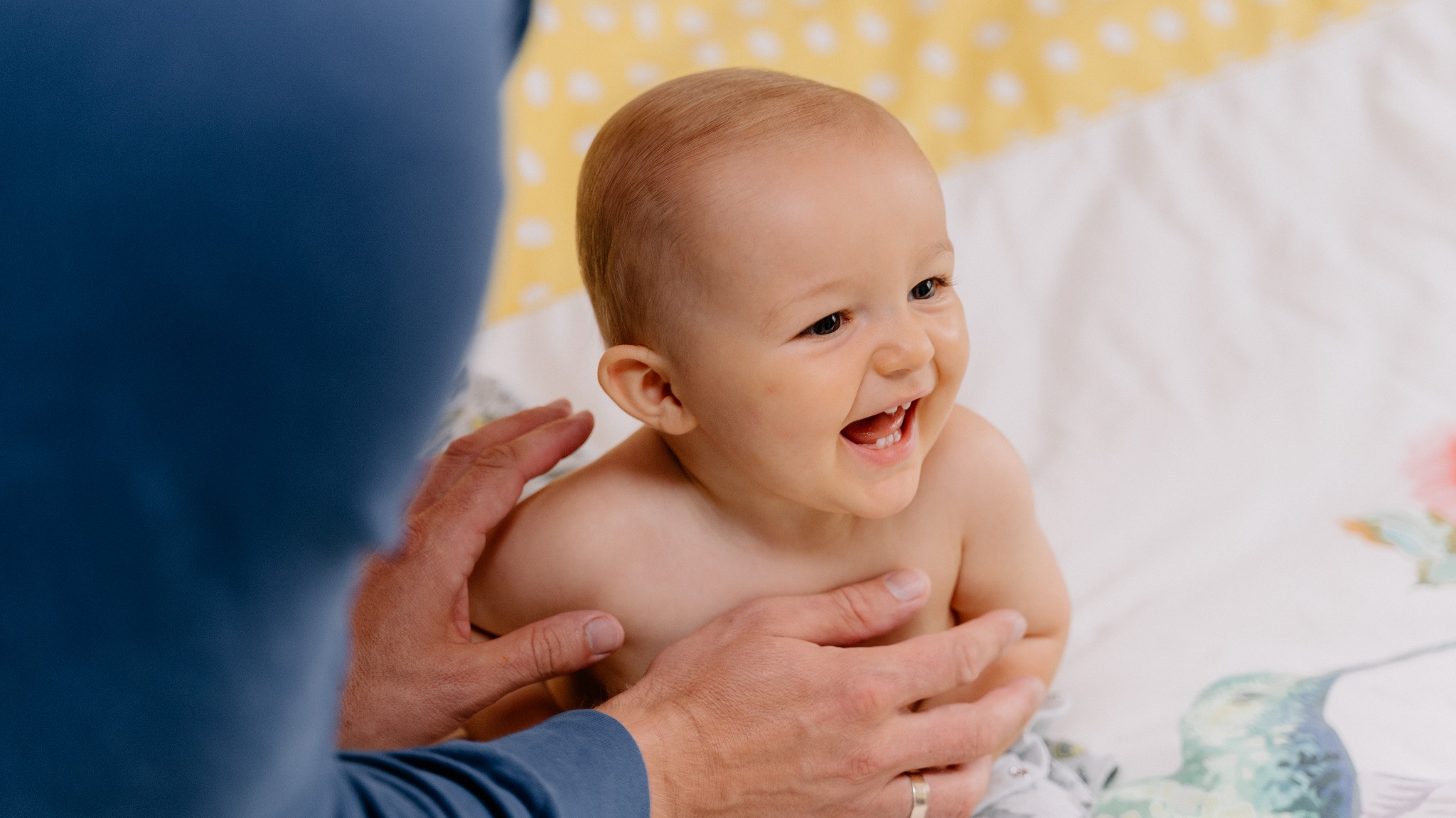 Uśmiechnięty niemowlak trzymany przez rodzica na wzorzystym kocyku z motywem kolibrów i kwiatów, widoczne dłonie rodzica z obrączką.