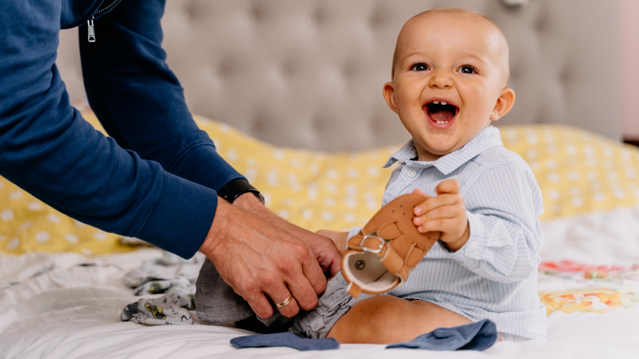 Uśmiechnięty niemowlak w błękitnej koszuli siedzi na łóżku, trzymając brązowy bucik w dłoni, podczas gdy dorosły pomaga mu założyć szare spodenki.
