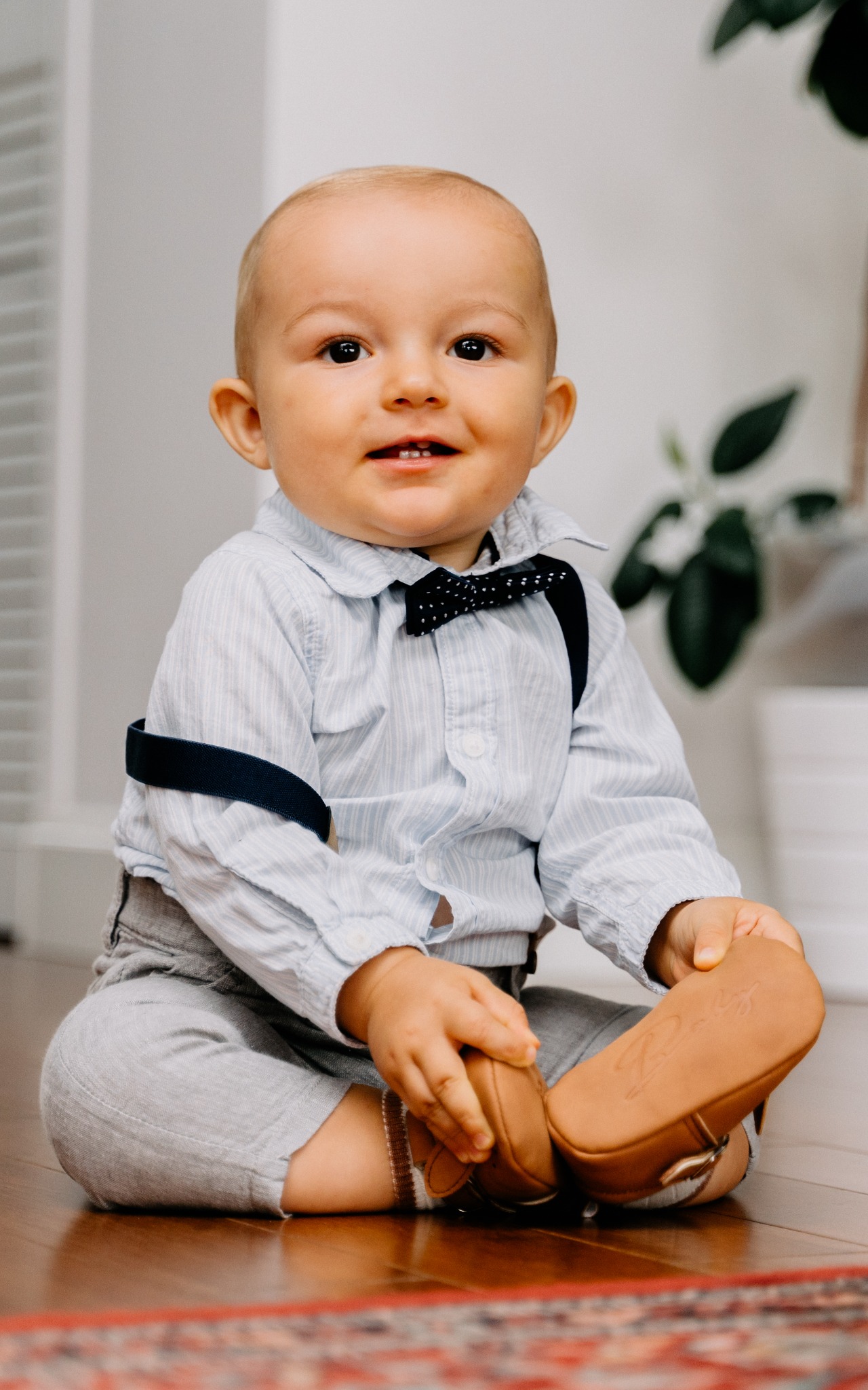 Uśmiechnięty niemowlak w niebieskiej koszuli w paski, z muszką i szelkami, siedzi na podłodze, trzymając w rączkach brązowe buciki.