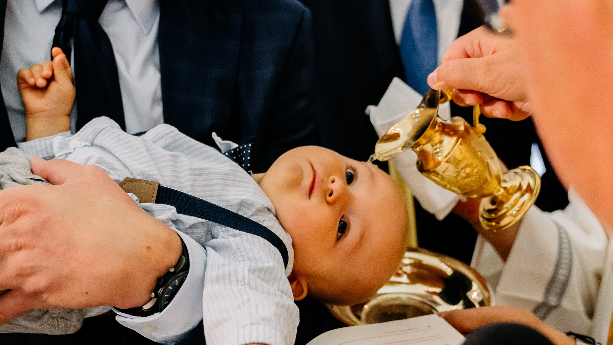Ujęcie z uroczystości chrztu: niemowlę w jasnej koszuli z szelkami spoczywa na rękach, podczas gdy z pozłacanej ampułki polewana jest woda.