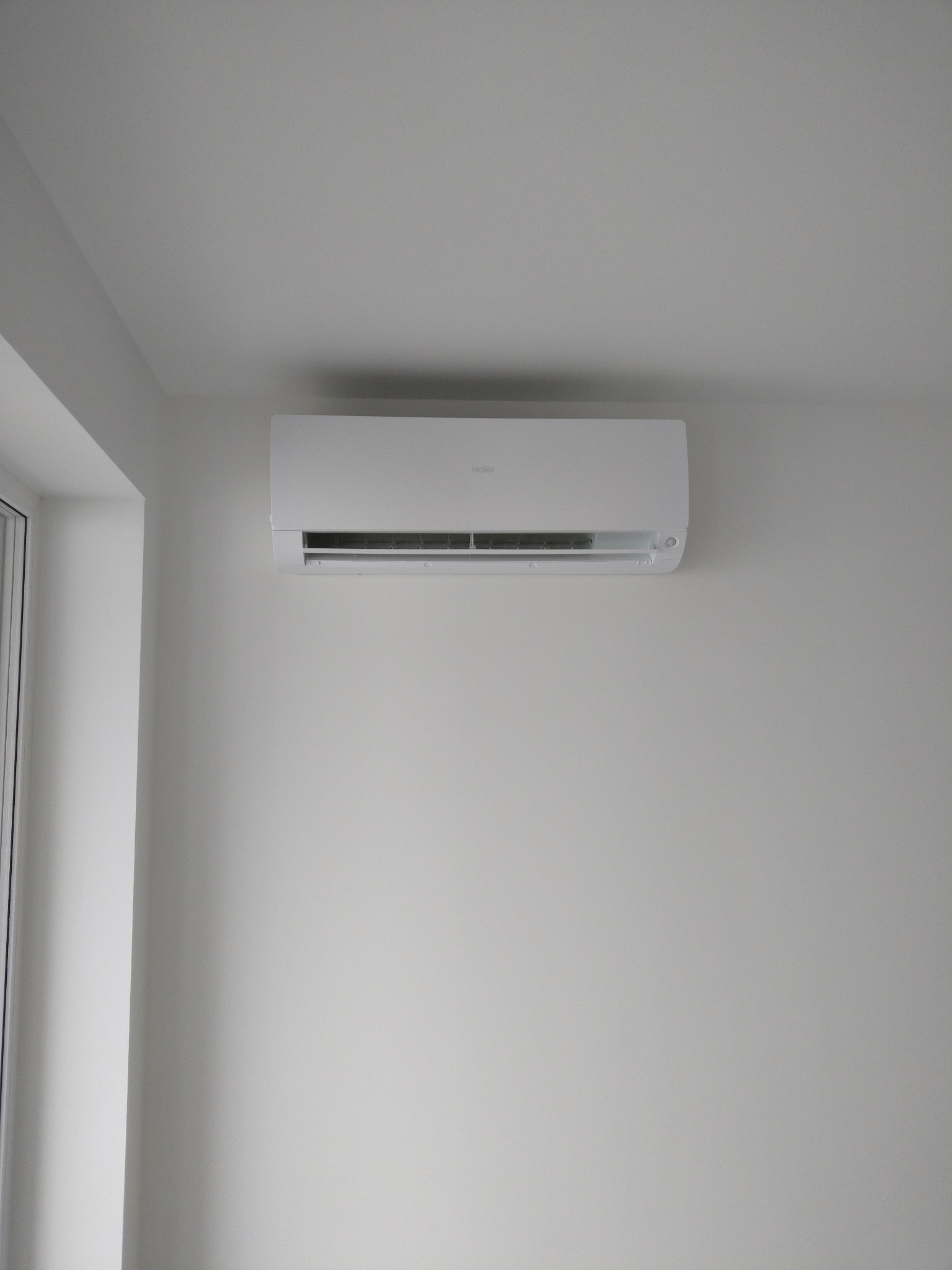 Biały klimatyzator marki Haier zamontowany na białej ścianie w minimalistycznym wnętrzu, widoczny fragment okna po lewej stronie.