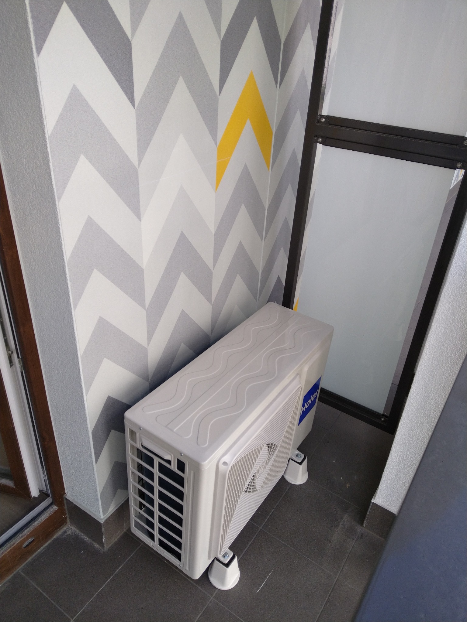 Biała jednostka zewnętrzna klimatyzatora Haier zamontowana na balkonie z szaro-białą tapetą w zygzaki i szarą podłogą z płytek.