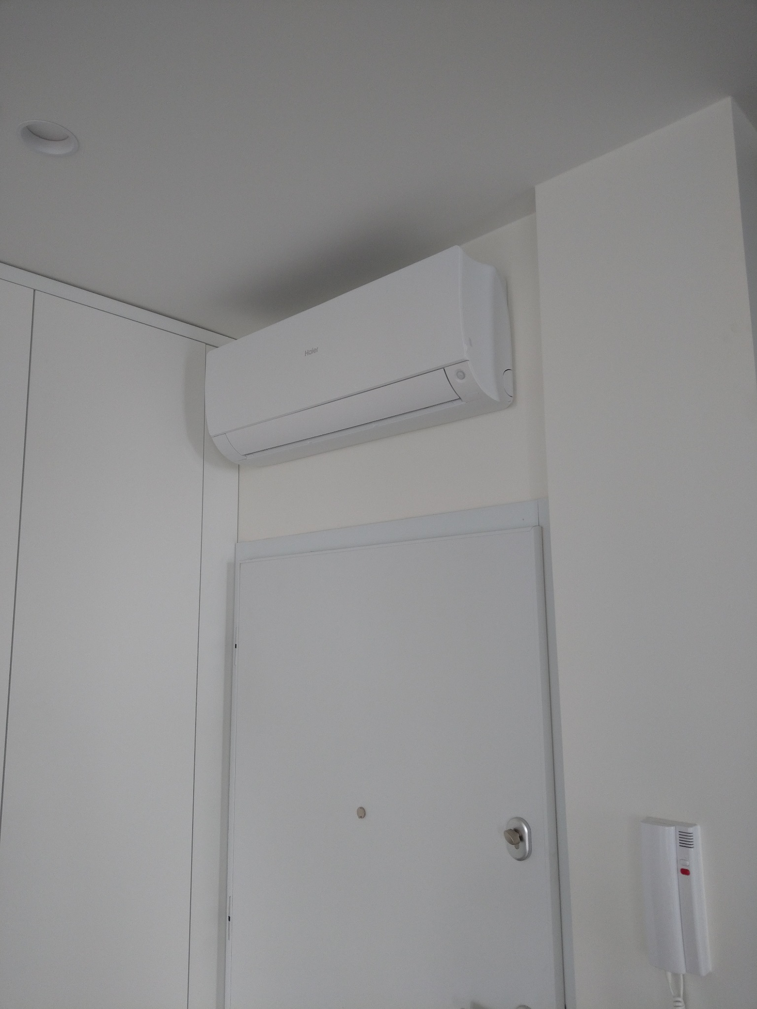 Biały klimatyzator marki Haier zamontowany nad drzwiami w jasnym pomieszczeniu, widoczny także domofon i szafa wnękowa.