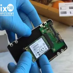 Technik w niebieskich rękawiczkach manipuluje dyskiem SSD M.2 w obudowie, widoczny fragment instrukcji i logo firmy serwisowej w rogu.