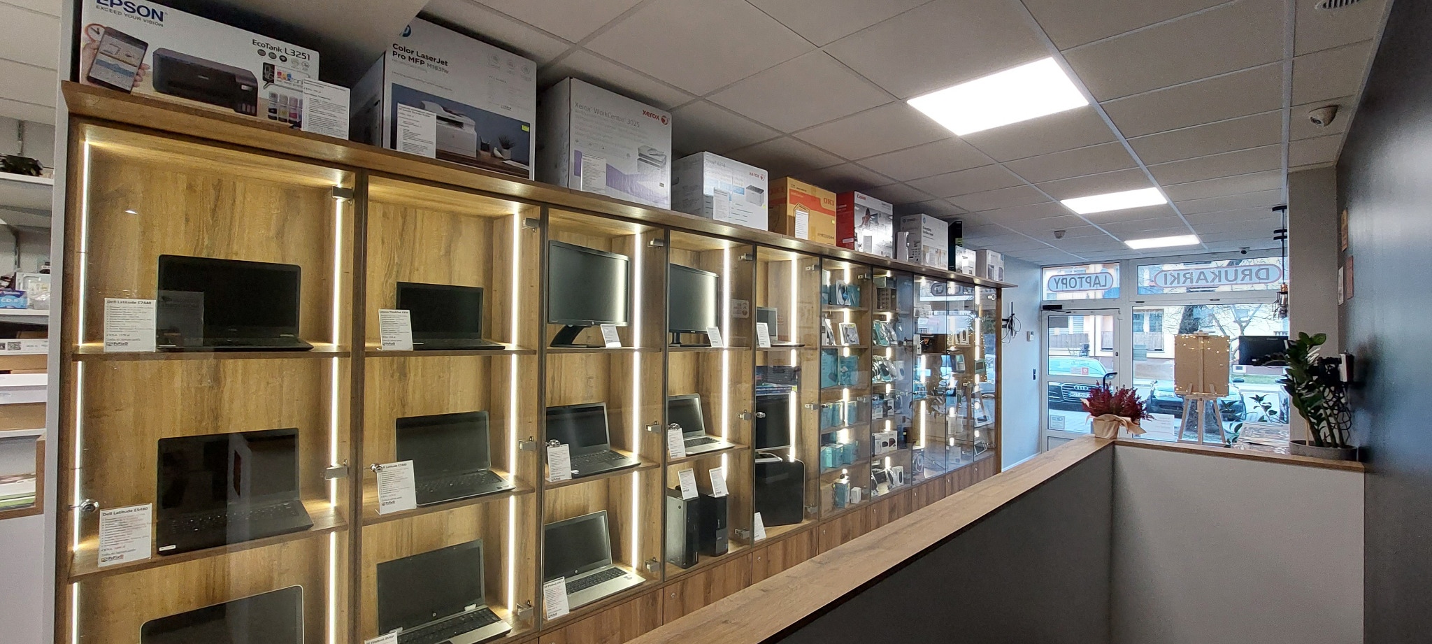 Wnętrze sklepu komputerowego z oświetlonymi półkami ekspozycyjnymi, na których prezentowane są laptopy, monitory i drukarki w pudełkach.