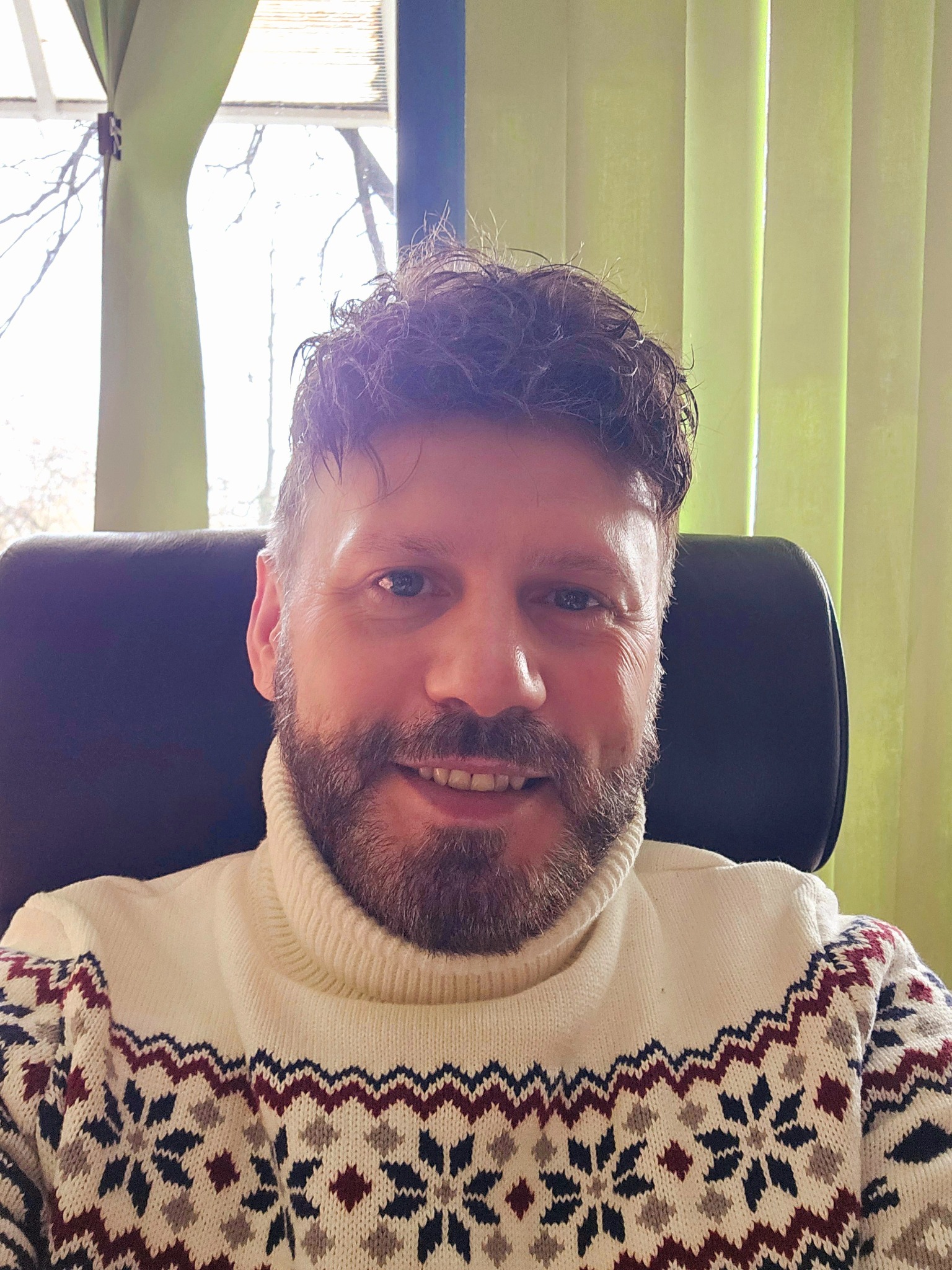 Portret uśmiechniętego mężczyzny z brodą, siedzącego w czarnym fotelu na tle okna z zielonymi żaluzjami, ubranego w kremowy sweter w norweskie wzory.