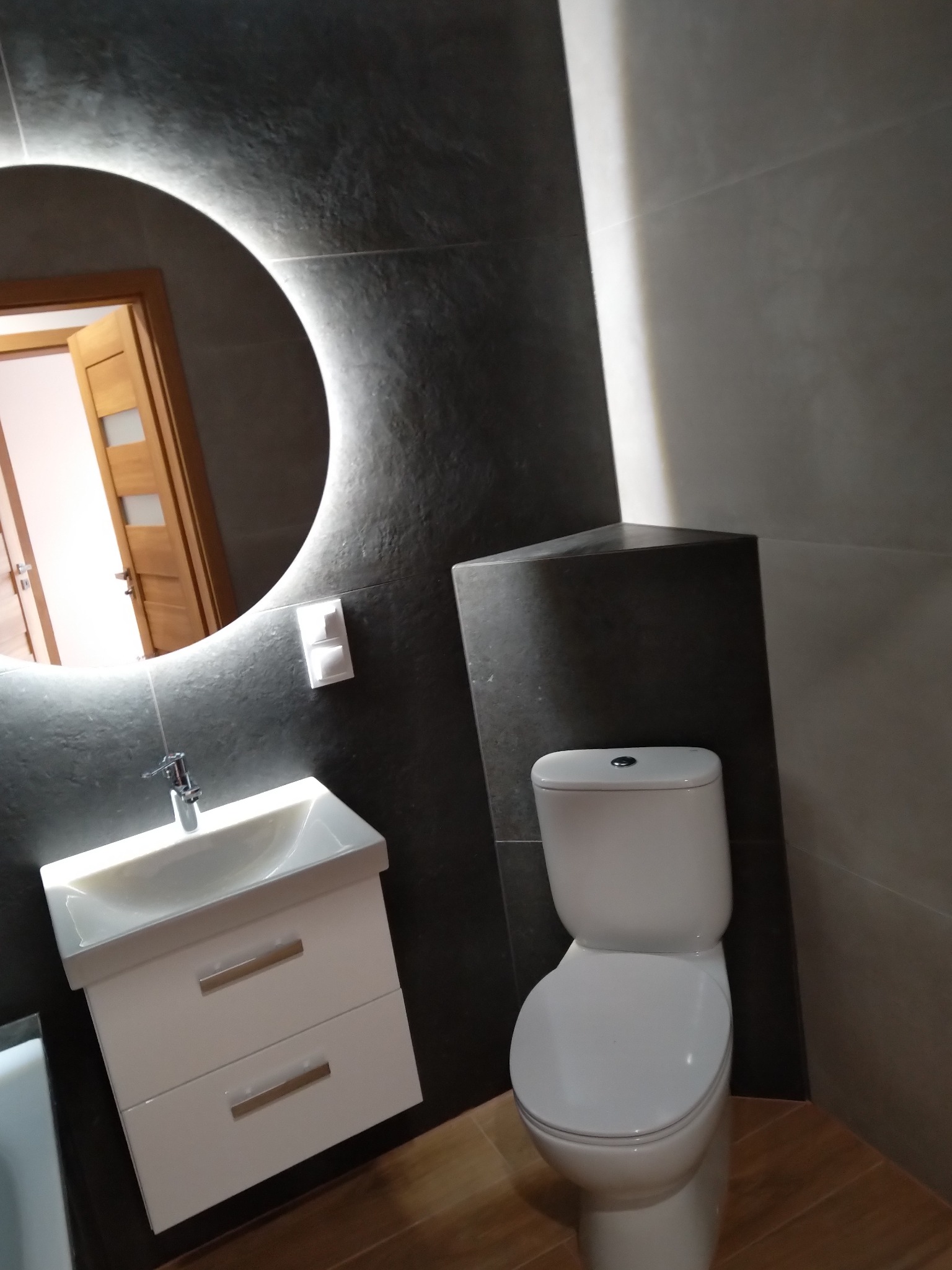 Nowoczesna łazienka z szarą płytką imitującą kamień, podświetlanym okrągłym lustrem, białą umywalką z szafką i wiszącą toaletą.