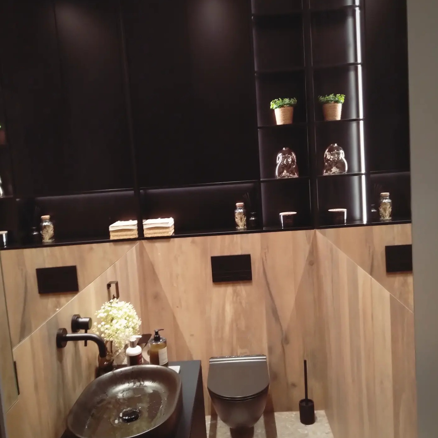 Nowoczesna łazienka z drewnopodobnymi płytkami na ścianach ułożonymi w geometryczny wzór, czarnymi szafkami wiszącymi z dekoracyjnymi elementami na półkach i owalną umywalką w kolorze ciemnego brązu.