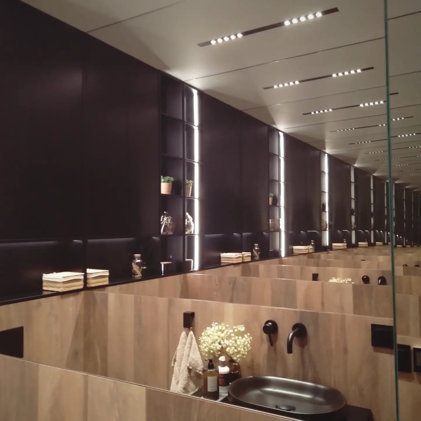 Nowoczesna łazienka z umywalkami w zabudowie z drewnopodobnych paneli, czarnymi bateriami i półkami z podświetleniem LED.