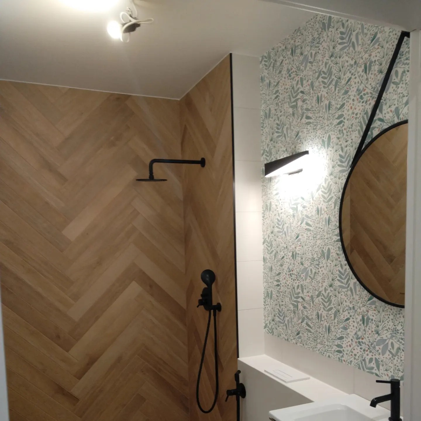 Nowoczesna łazienka z drewnianą ścianą prysznicową ułożoną w jodełkę, czarną armaturą i ścianą pokrytą tapetą w roślinny wzór, okrągłe lustro z czarną ramą.