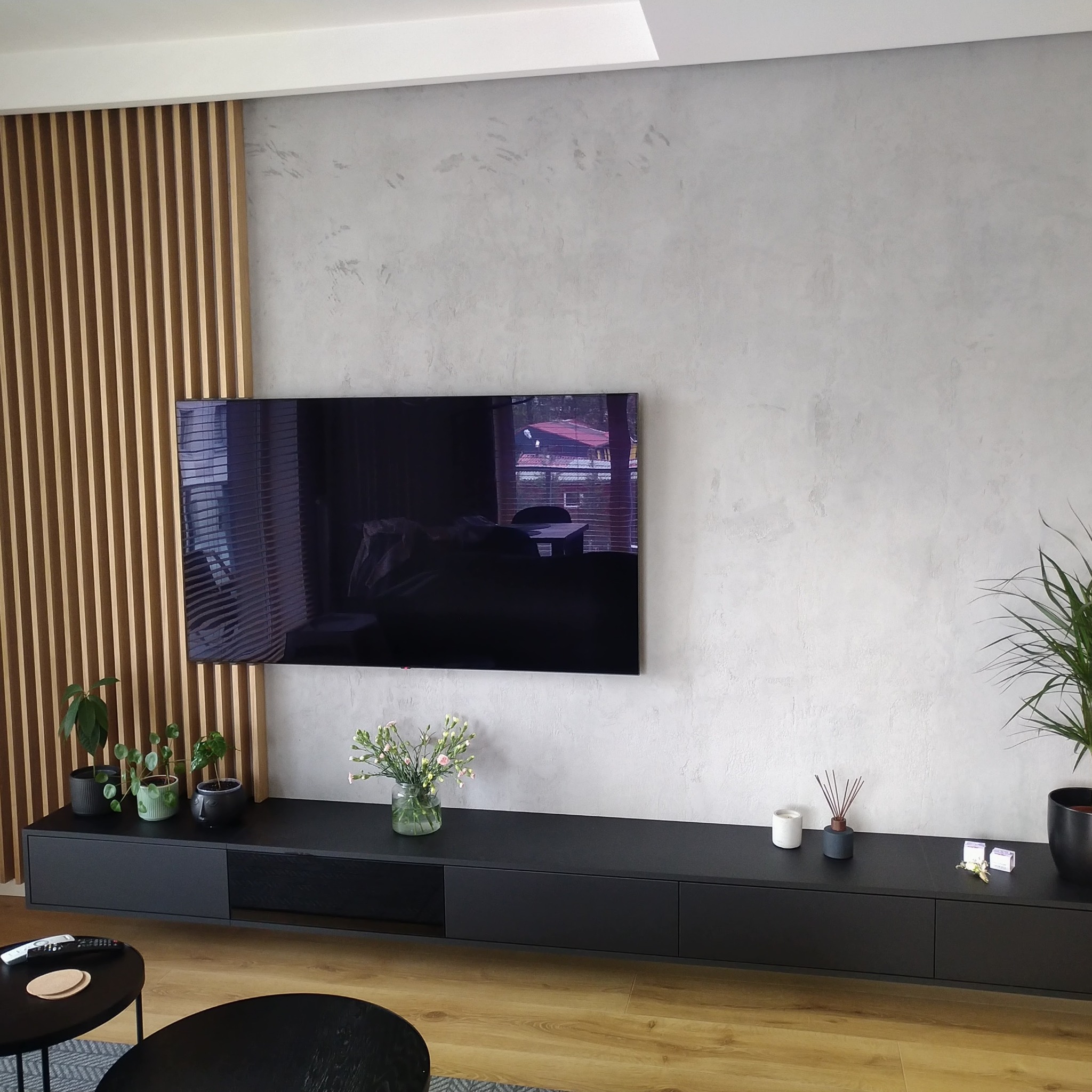 Minimalistyczny salon z szarą ścianą o fakturze betonu, drewnianymi lamelami dekoracyjnymi, zawieszonym czarnym telewizorem i szafką RTV, podkreślony przez rośliny doniczkowe i drewnianą podłogę.