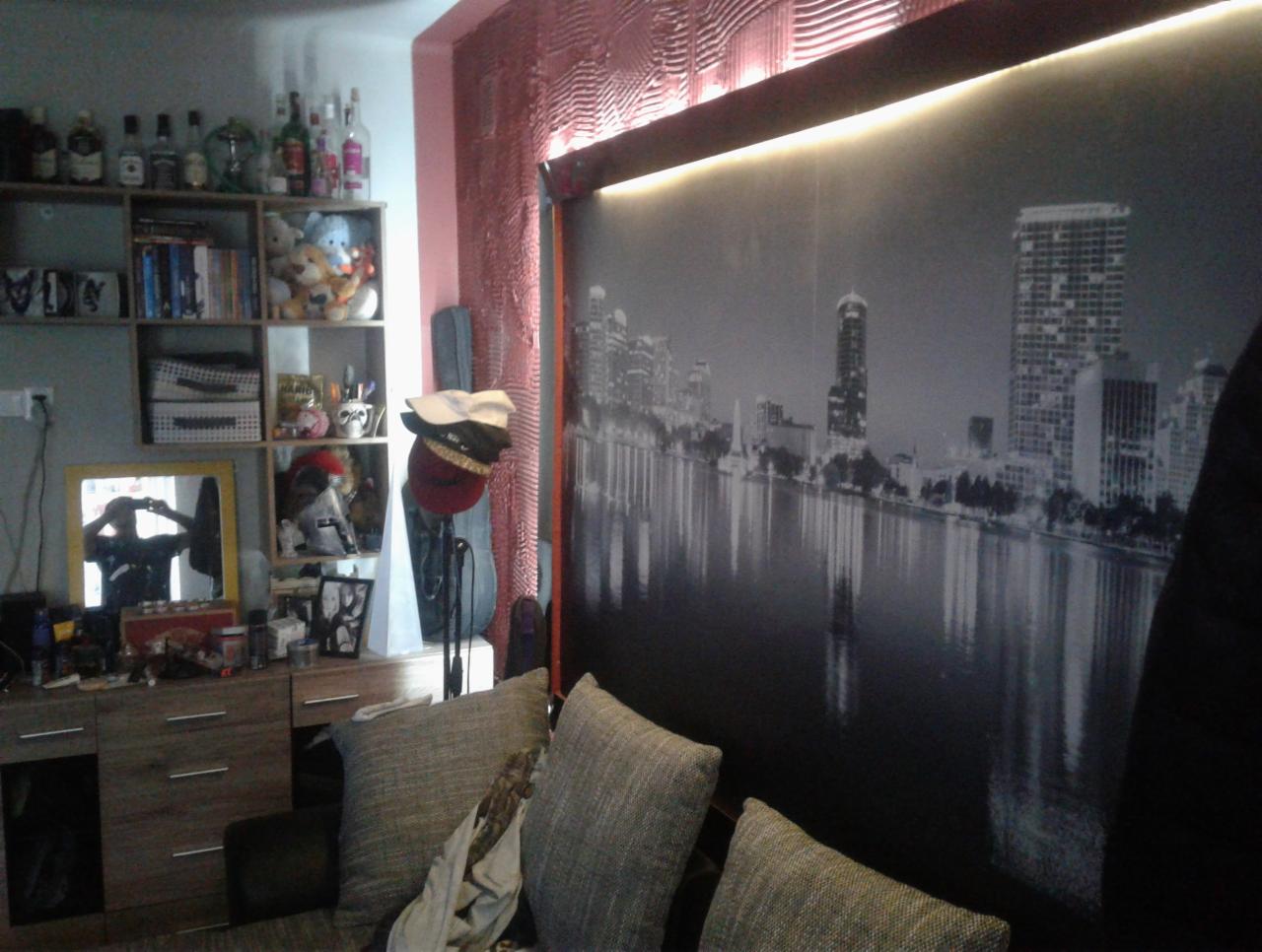 Fragment pokoju z fototapetą przedstawiającą panoramę miasta nad wodą, widoczna część sofy z poduszkami, komoda z półkami i różnymi przedmiotami, w tym lustro z odbiciem osoby robiącej zdjęcie.