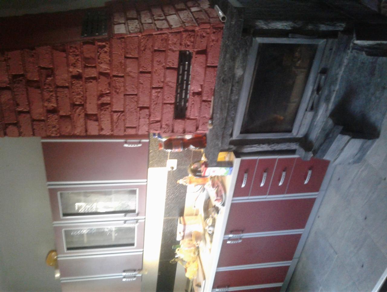 Część kuchni z zabudową meblową w odcieniach bordo i ceglaną ścianą z wnęką, nad blatem kuchennym widoczne przyprawy i akcesoria.