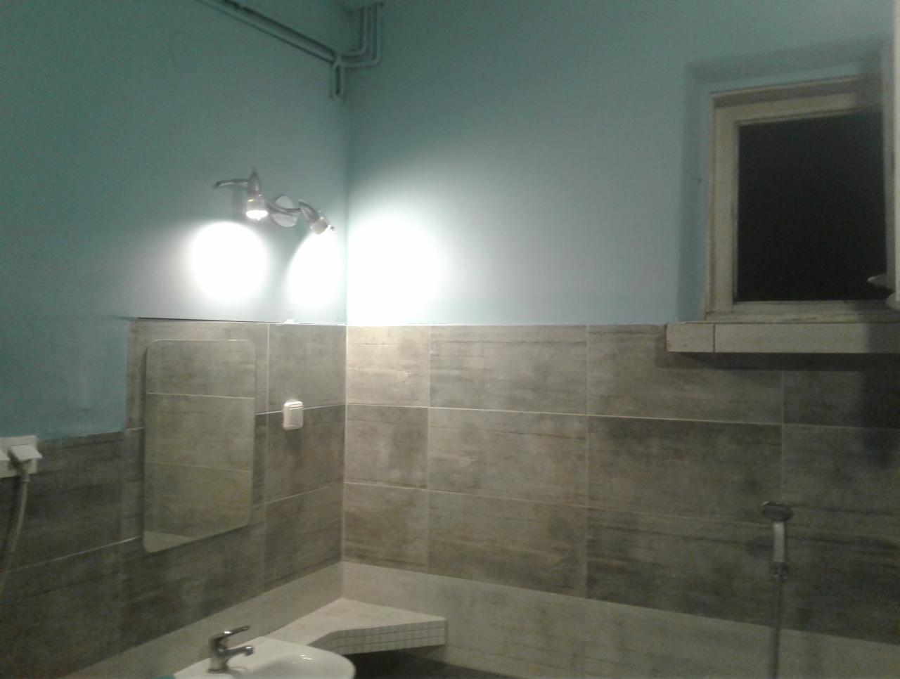 Szara łazienka z niebieskimi ścianami, widoczne płytki na ścianach, umywalka i okno z czarnym tłem, oświetlenie sufitowe.