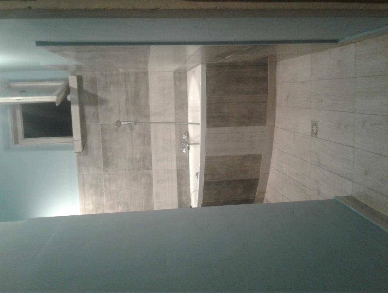 Wykończona łazienka z kabiną prysznicową typu walk-in, wyłożona płytkami w odcieniach szarości i beżu, z odpływem liniowym w podłodze i oknem dachowym.