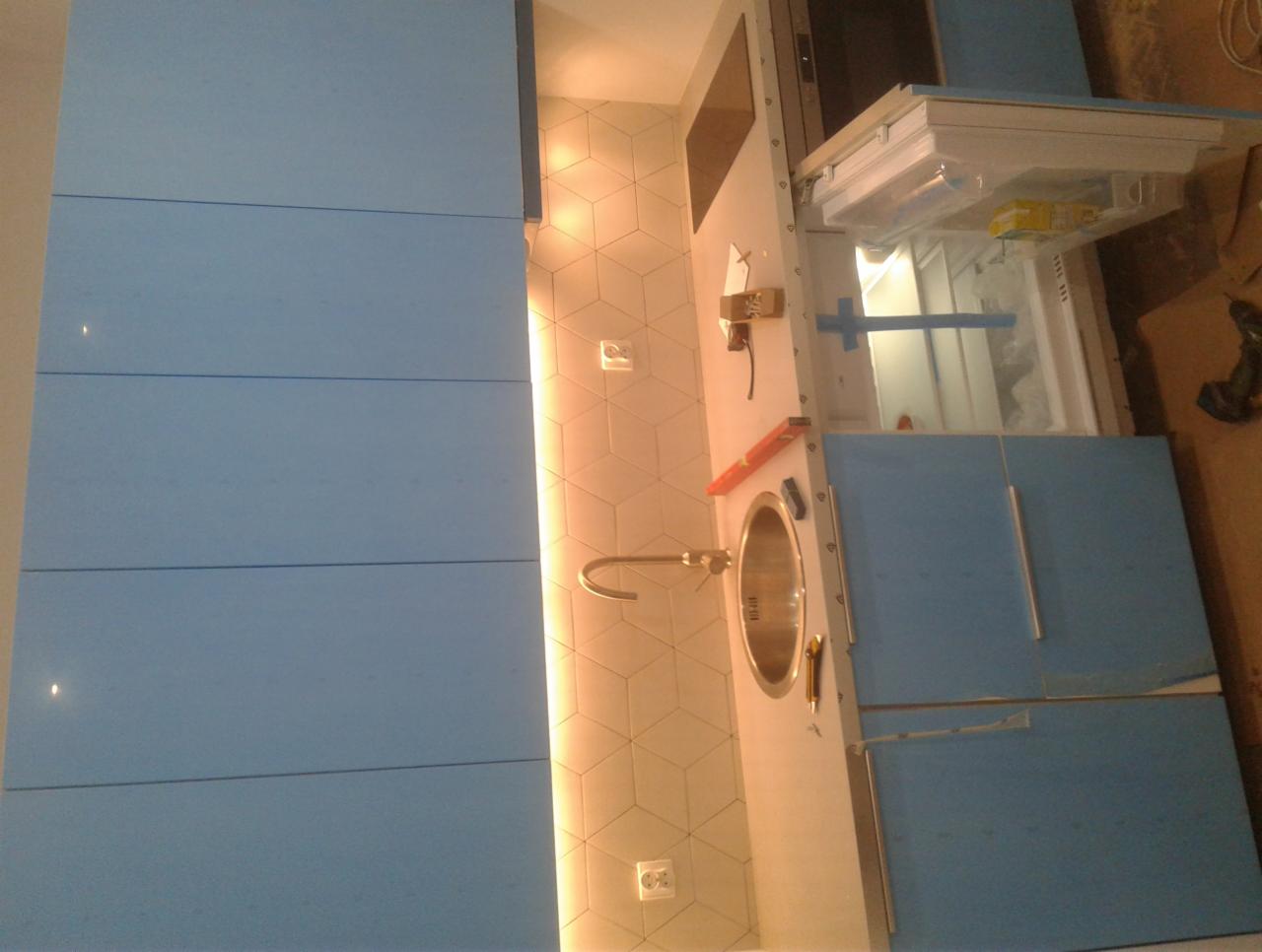 Niebieska zabudowa kuchenna z oświetleniem LED na ścianie z geometrycznymi płytkami, widoczny zlew z baterią oraz otwarta lodówka w trakcie montażu.