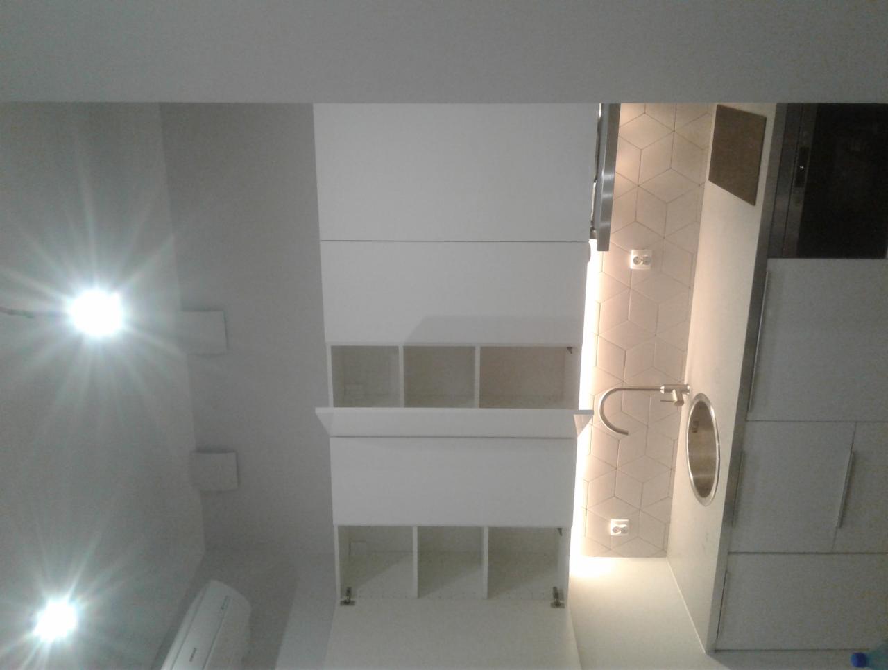 Nowoczesna, biała łazienka z szafkami wiszącymi, umywalką wpuszczaną w blat i heksagonalnymi płytkami na ścianie, oświetlona punktowo.