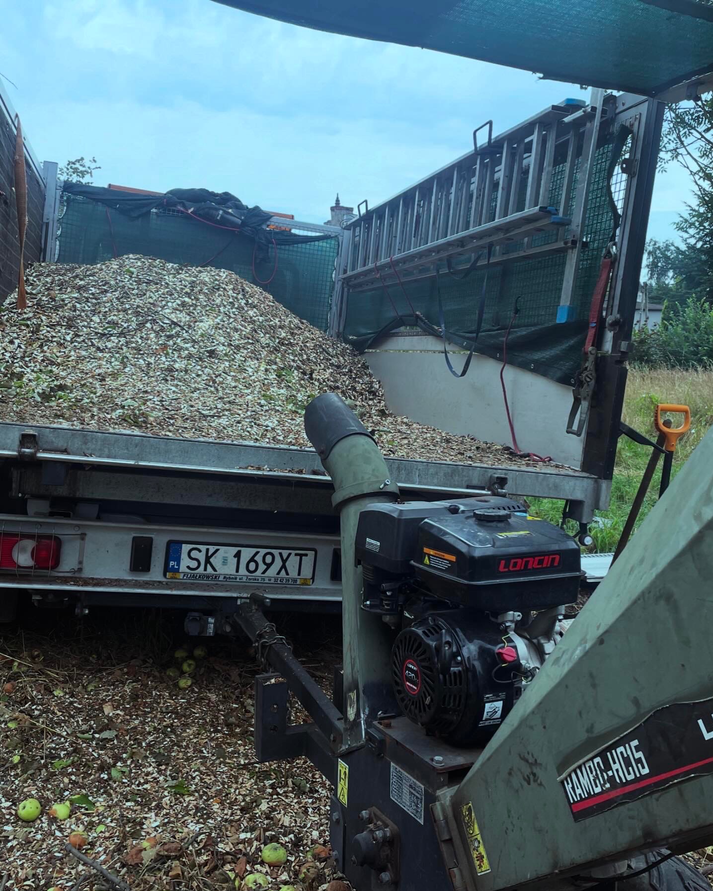 Przyczepa wypełniona zrębkami drzewnymi po wycince, obok rozdrabniacz do gałęzi marki Loncin, widoczny numer rejestracyjny SK 169XT.