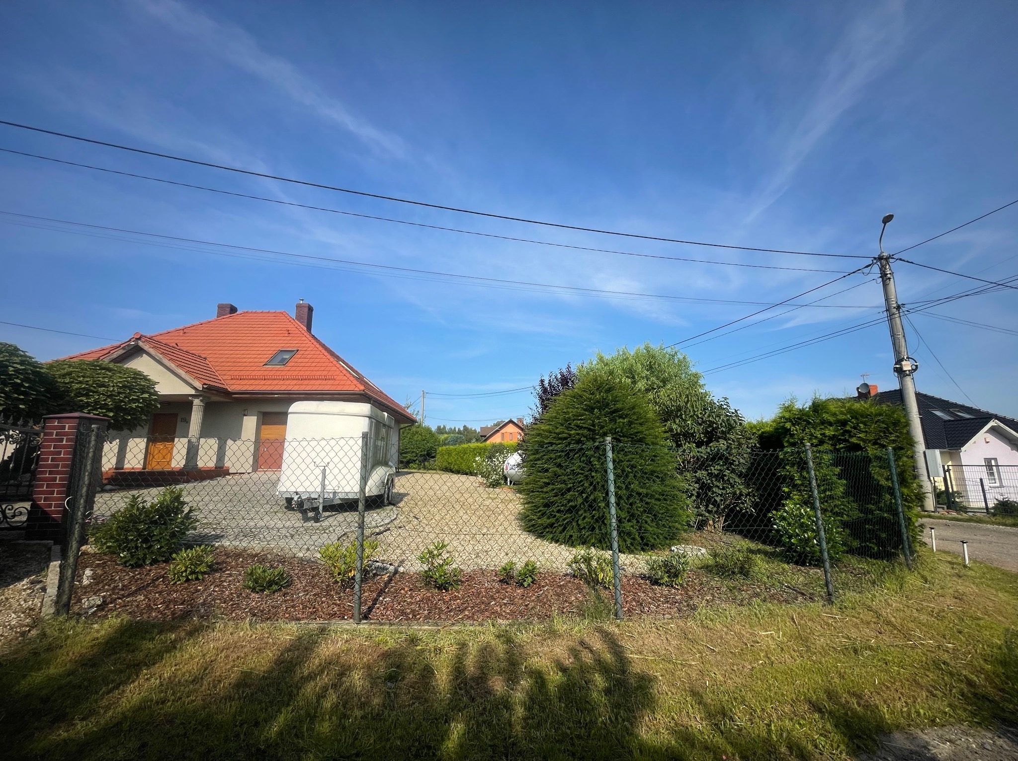 Ogród z niskim metalowym ogrodzeniem, obsadzony krzewami i korą, na tle domu z czerwoną dachówką i błękitnego nieba z chmurami.