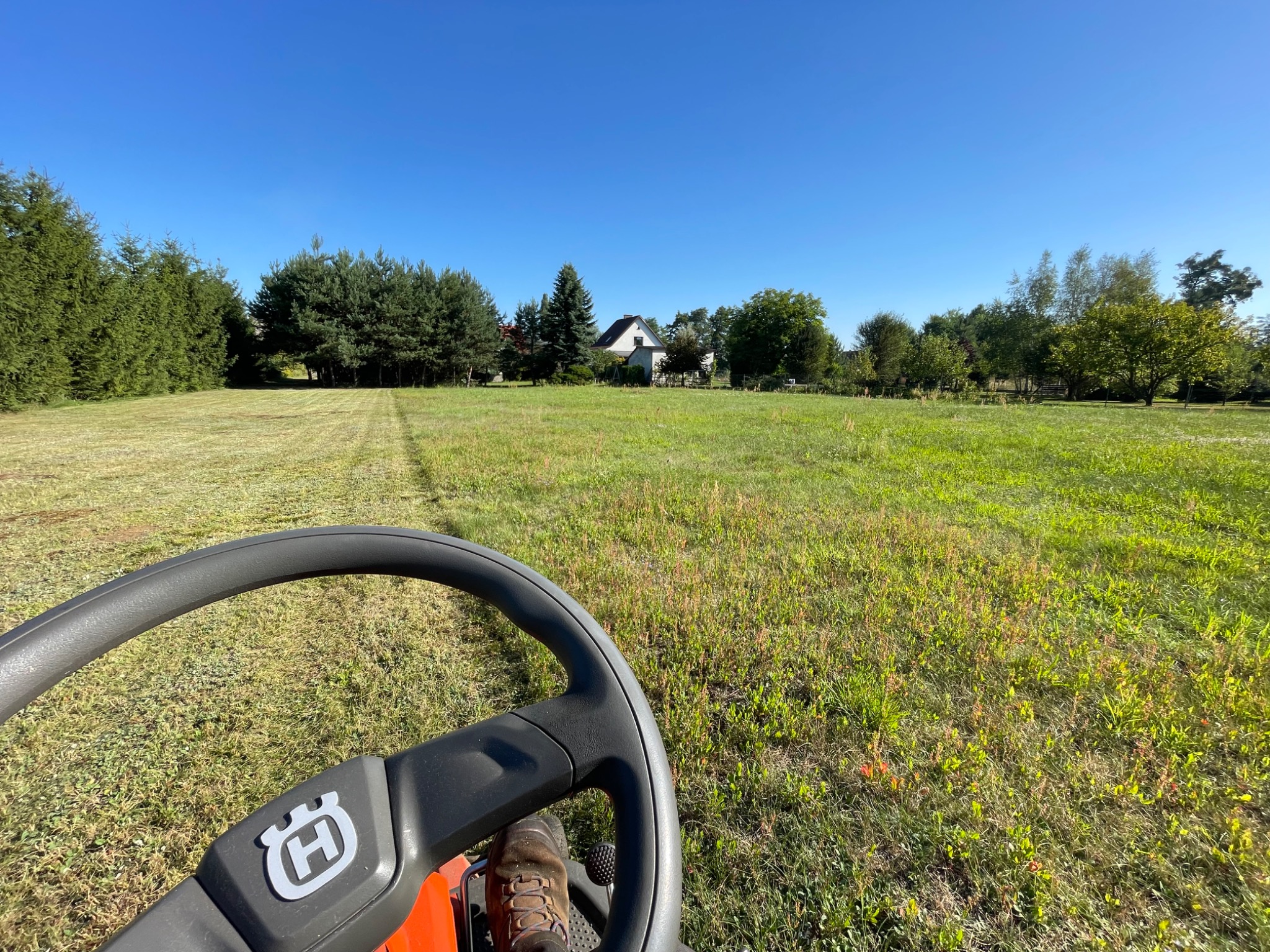 Widok z traktorka Husqvarna na skoszone pole, gdzie w tle widoczny jest dom i drzewa, a na pierwszym planie kierownica i but operatora.