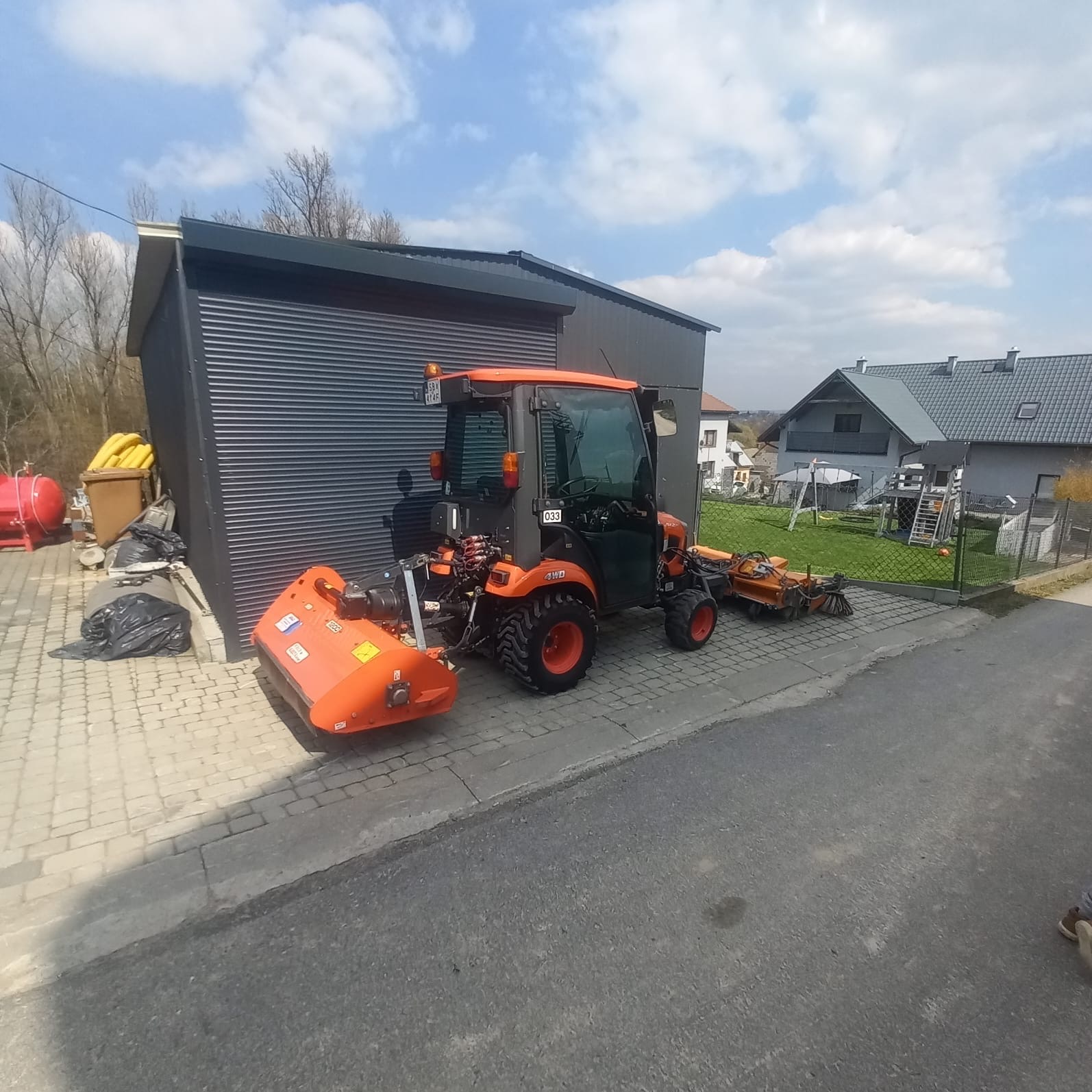 Pomarańczowy traktor z kosiarką i zamiatarką na kostce brukowej, na tle szarego garażu i domu jednorodzinnego w słoneczny dzień. Widoczne drzewa bez liści.