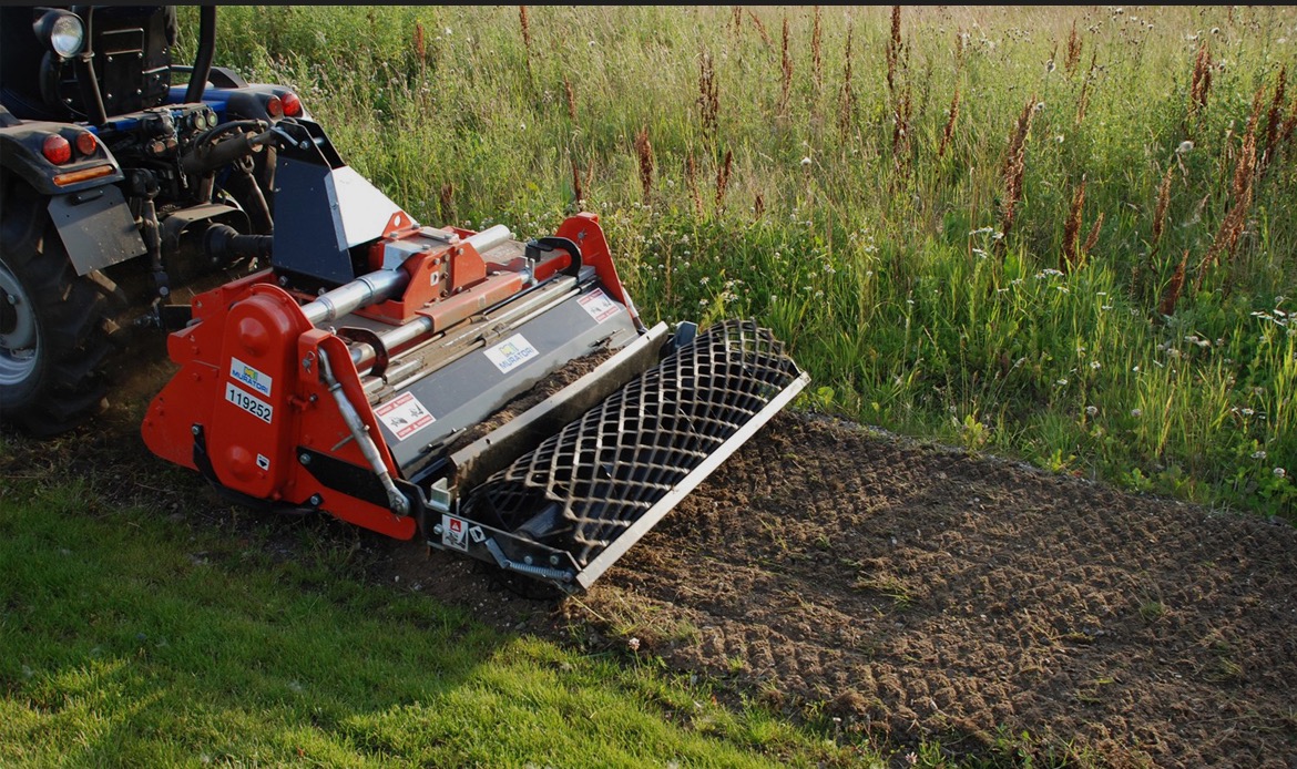 Czerwony traktor z glebogryzarką Muratori spulchnia ziemię na polu, przygotowując ją pod uprawę. Widoczne ślady pracy maszyny na tle zielonej trawy i roślinności.
