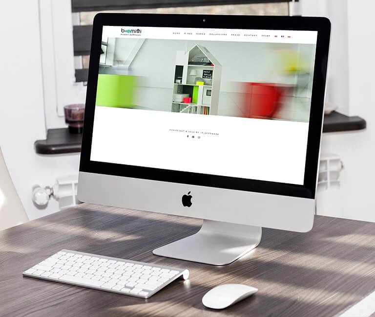 Ekran komputera iMac prezentujący stronę internetową z minimalistycznym designem i zdjęciem regału w kształcie domu, na biurku leży bezprzewodowa klawiatura i mysz Apple.