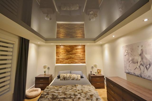 Nowoczesna sypialnia z drewnianą dekoracją ścienną i błyszczącym, podwieszanym sufitem, odbijającym elementy wystroju, uzupełniona obrazem koni i minimalistycznymi lampkami nocnymi.