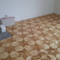 Układanie parkietu w geometryczny wzór 3D w jasnym pomieszczeniu, widoczna część podłogi z szarymi płytami oraz pojemnik z klejem.