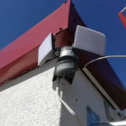 Zewnętrzna lampa LED z czujnikiem ruchu zamontowana na elewacji budynku pod czerwonym okapem, widok z dołu na tle błękitnego nieba.