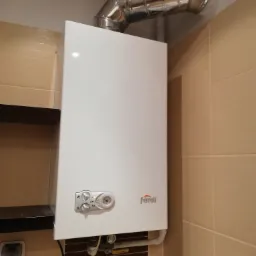 Biały, wiszący piec gazowy marki Ferroli z chromowaną rurą odprowadzającą spaliny, zamontowany na ścianie wyłożonej beżowymi płytkami ceramicznymi.