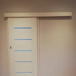 Białe drzwi przesuwne z poziomymi, błękitnymi wstawkami, zamontowane na szarej ścianie, widoczne z bliska, z drewnianą podłogą.