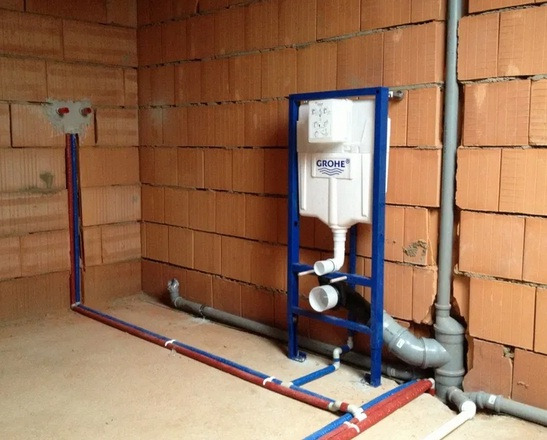 Stelaż podtynkowy Grohe zamontowany na tle surowej ściany z cegły, widoczne rury instalacji wodnej i kanalizacyjnej w trakcie montażu.