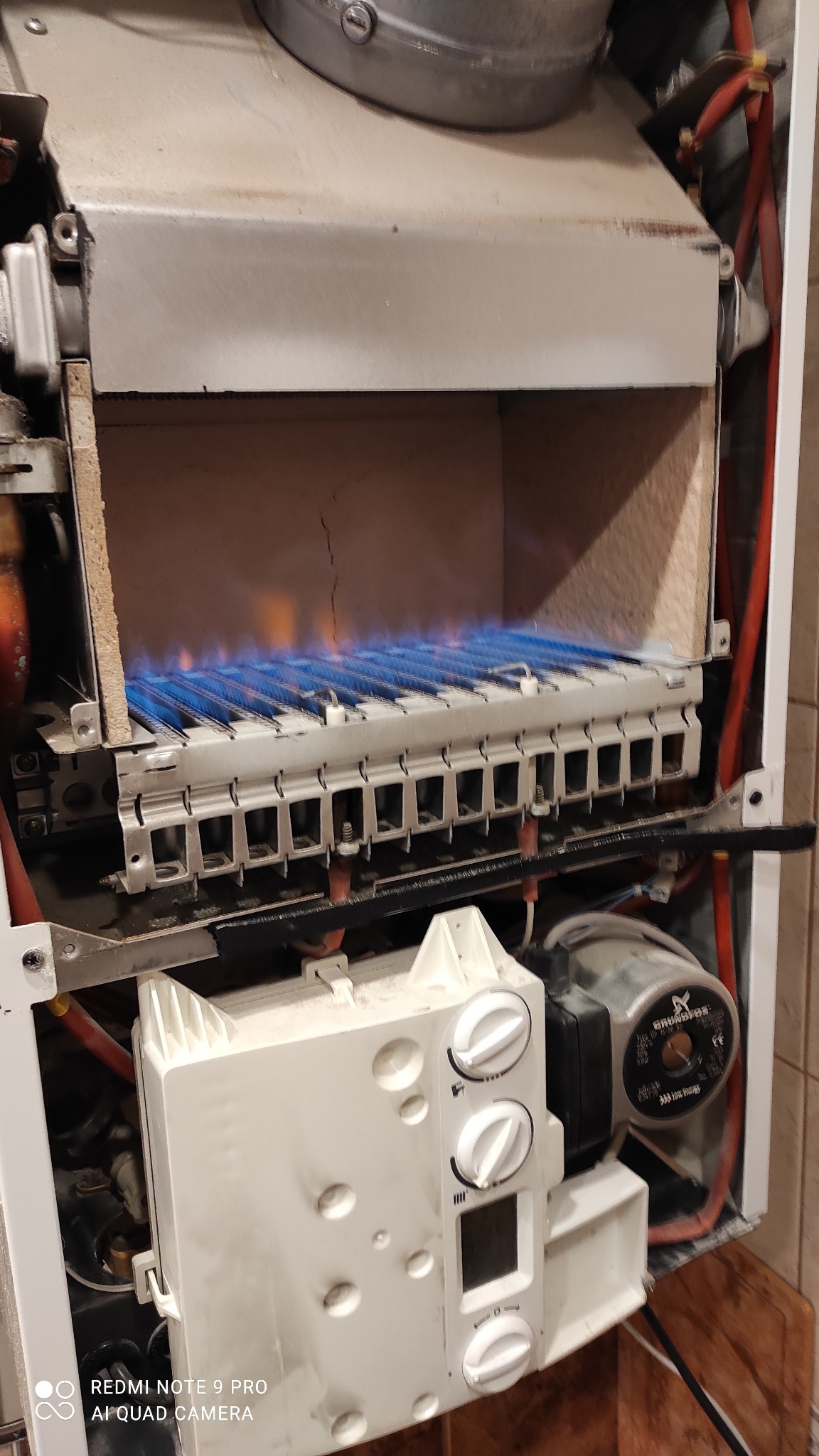 Otwarty piec gazowy z widocznymi płomieniami palnika, biała obudowa z pokrętłami i wyświetlaczem, widoczne elementy instalacji.