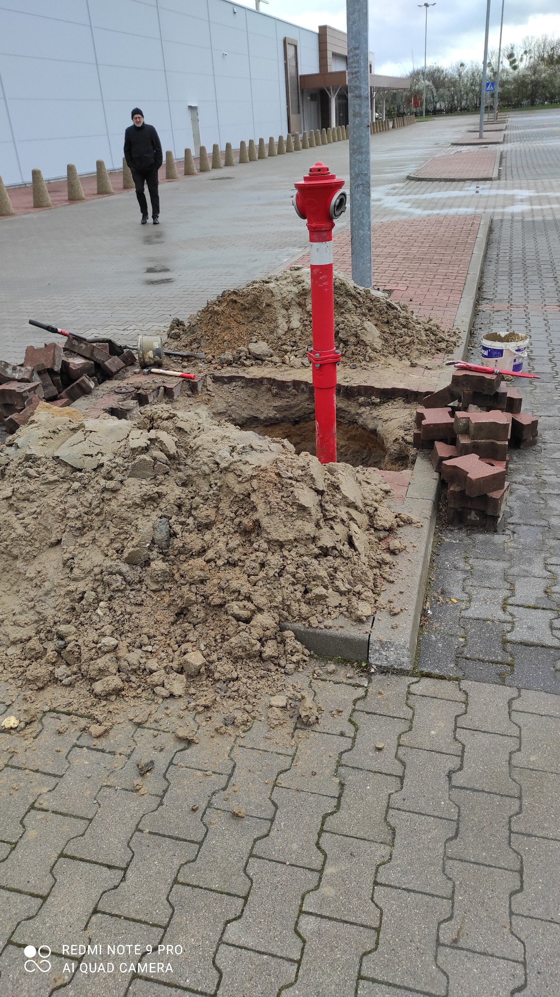 Czerwony hydrant wystający z wykopu, obok sterta ziemi, kostka brukowa i porozrzucane cegły, w tle budynek i przechodzień.