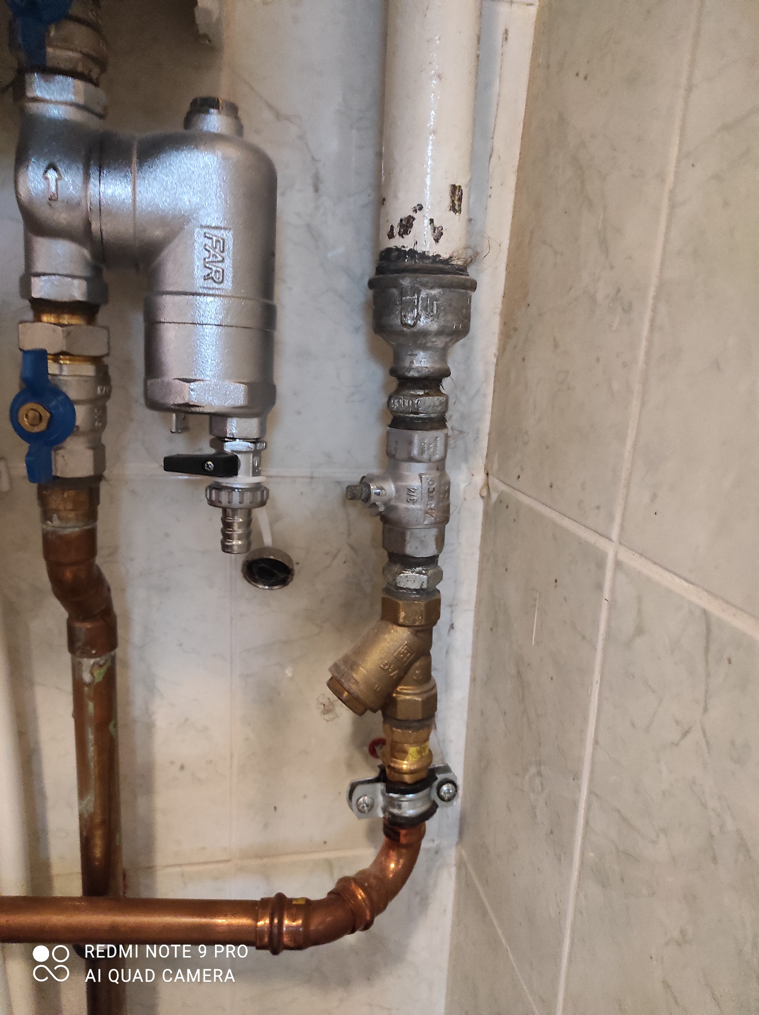 Fragment instalacji hydraulicznej z rur miedzianych, zaworów i filtra FAR na tle marmurowej ściany.