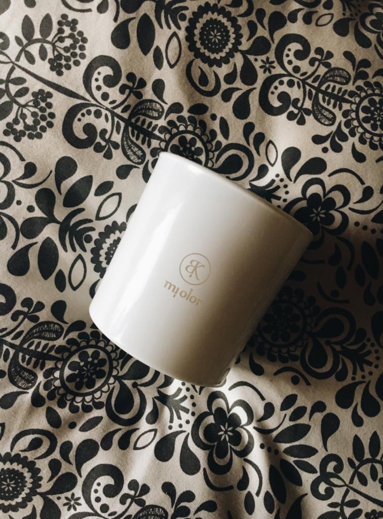 Biała świeca zapachowa 'mIolor' z delikatnym złotym logo na wzorzystej, czarno-białej tkaninie w kwiaty.