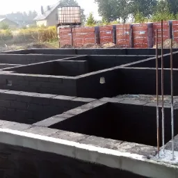 Wznoszenie fundamentów z bloczków Termoton, zabezpieczonych czarną izolacją, widoczne wystające pręty zbrojeniowe i palety z bloczkami w tle.