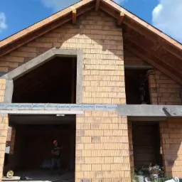 Wznoszenie dwukondygnacyjnego budynku z pustak&oacute;w ceramicznych; widoczne otwory okienne i drzwiowe; drewniana konstrukcja dachu w trakcie montażu; pracownik wewnątrz budynku.