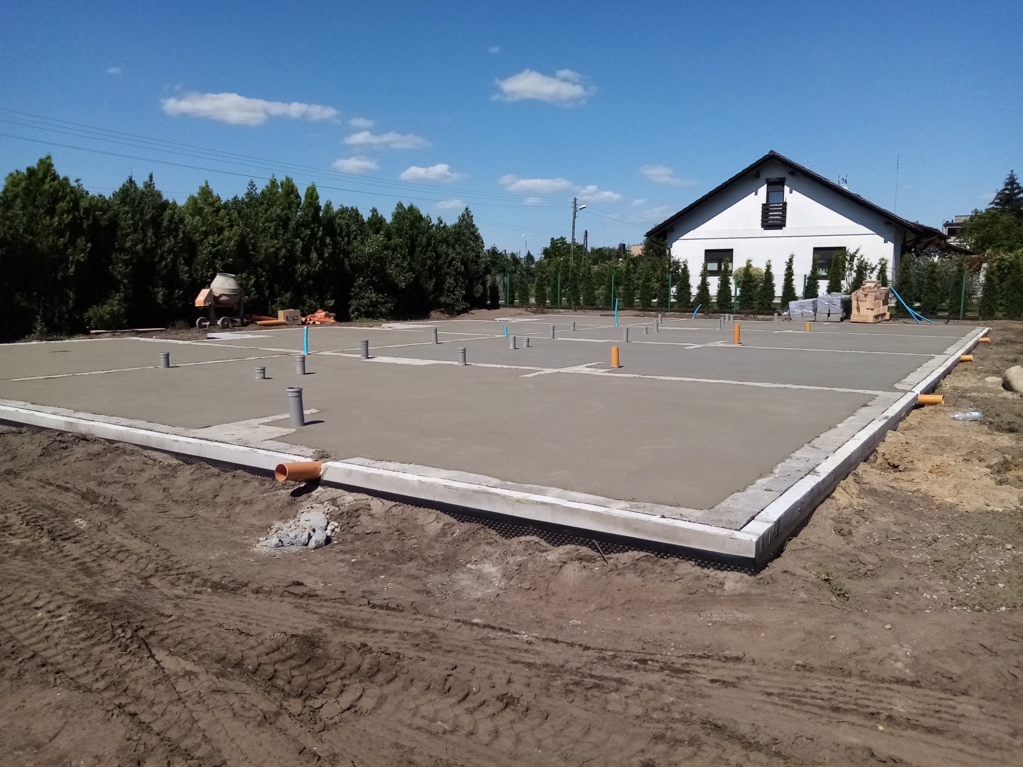 Wylany fundament pod budowę domu jednorodzinnego z widocznymi pionowymi rurami instalacyjnymi i betonową obwódką, w tle dom w stanie surowym otoczony zielenią.