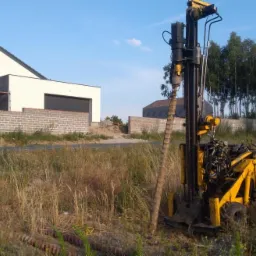 Żółta wiertnica geologiczna na tle budowy domu jednorodzinnego z szarej cegły, w oddali widoczny las.