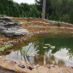 Oczko wodne z kaskadą skalną, obłożone kamieniami, roślinnością wodną i otoczone iglakami; robocze rękawice leżą na pierwszym planie.